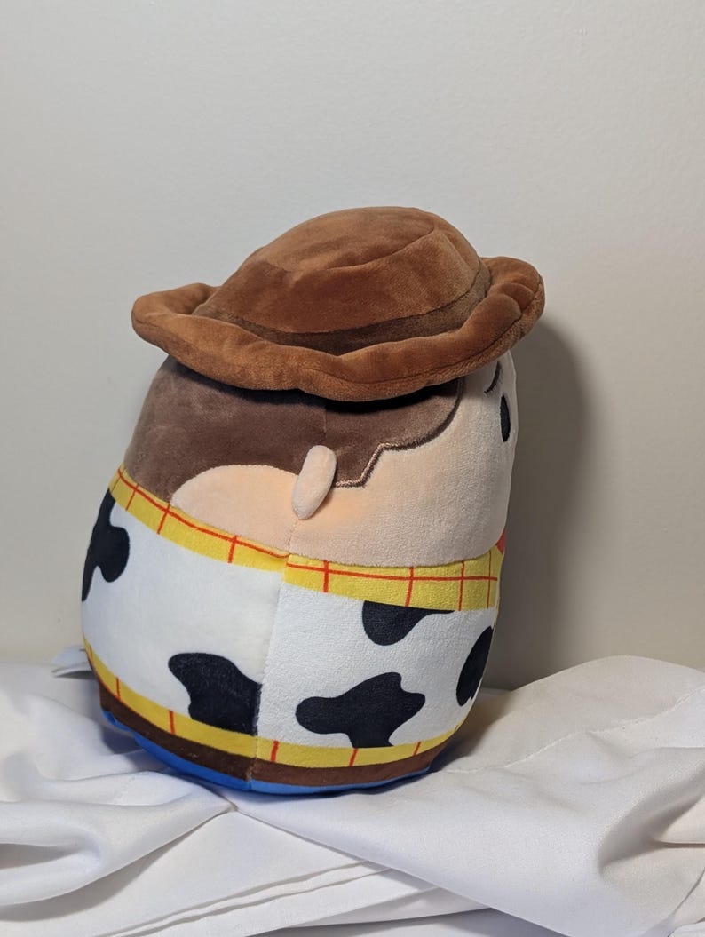 May include: A plush toy of the character Woody from the movie Toy Story. The toy is brown, tan, and white with black spots, and a brown cowboy hat. The toy has a blue base and yellow trim.