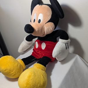 May include: A plush Mickey Mouse doll, featuring a black head with large ears, a tan face, and a black nose. It wears red shorts, yellow shoes, and white gloves. The doll is soft and cuddly, perfect for children and Disney fans.