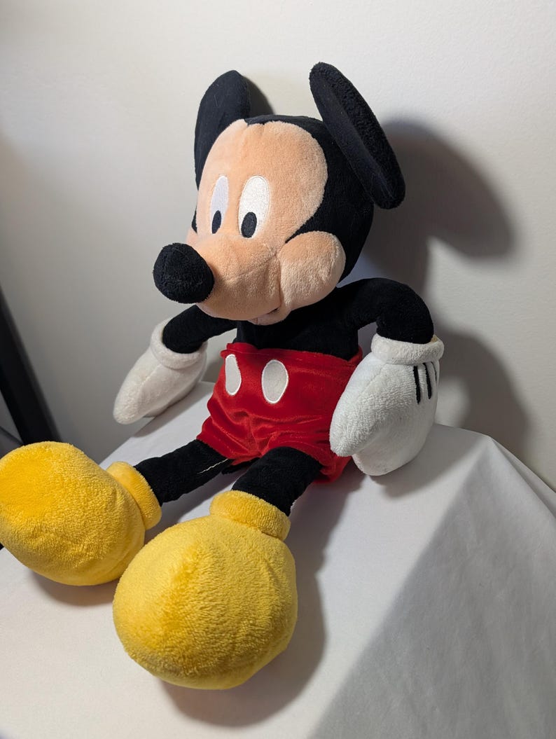 May include: A plush Mickey Mouse doll, featuring a black head with large ears, a tan face, and a black nose. It wears red shorts, yellow shoes, and white gloves. The doll is soft and cuddly, perfect for children and Disney fans.