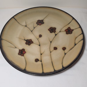 May include: A round ceramic plate featuring a beige base color with dark brown, segmented branch-like patterns and small, dark red floral accents. The plate has a smooth, matte finish and a dark brown rim that frames the overall design.