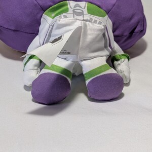 May include: A plush Buzz Lightyear doll, viewed from the back, showcasing its signature white and green spacesuit design. The doll features purple accents on the feet and a large purple head. A tag is visible on the back of the doll.
