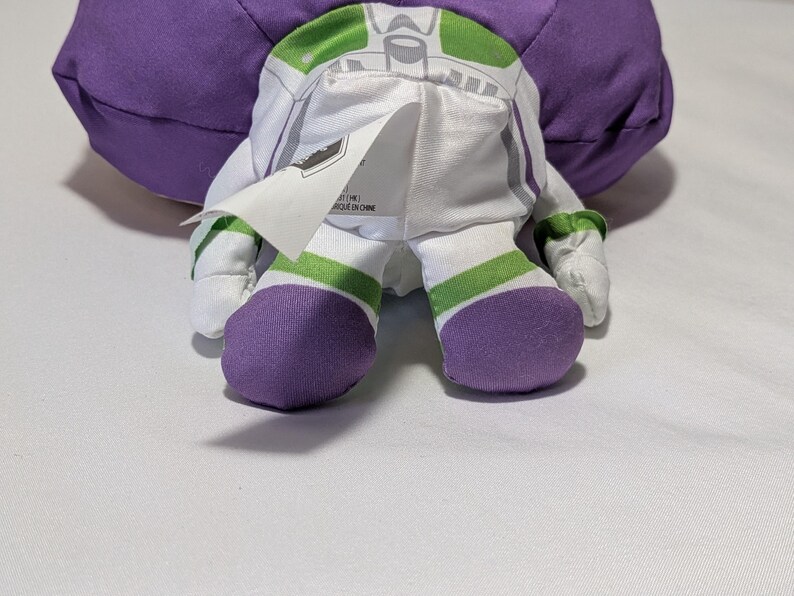 May include: A plush Buzz Lightyear doll, viewed from the back, showcasing its signature white and green spacesuit design. The doll features purple accents on the feet and a large purple head. A tag is visible on the back of the doll.