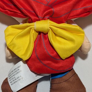 May include: A soft toy doll featuring a red outfit and a large yellow bow. The doll has brown boots and a tag with text. The bow is centrally placed on the doll's back, with a vibrant and colourful design.