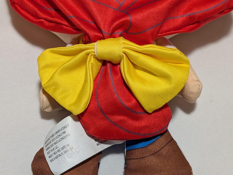 May include: A soft toy doll featuring a red outfit and a large yellow bow. The doll has brown boots and a tag with text. The bow is centrally placed on the doll's back, with a vibrant and colourful design.