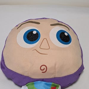 May include: A plush toy featuring the face of Buzz Lightyear from Toy Story. The toy has a light tan face with large blue eyes, a small nose, and a smiling mouth. The head is framed by purple fabric.