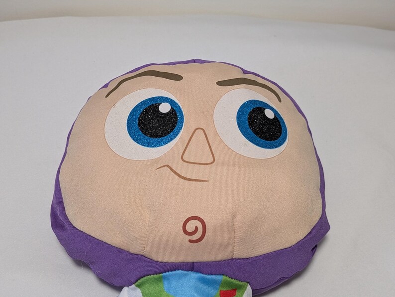 May include: A plush toy featuring the face of Buzz Lightyear from Toy Story. The toy has a light tan face with large blue eyes, a small nose, and a smiling mouth. The head is framed by purple fabric.