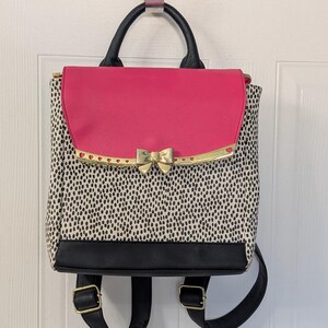 May include: A backpack featuring a black and white dalmatian-style spotted pattern, a solid hot pink top flap, and a black base. The bag includes a gold-toned metal bow accent on the front flap, a black top handle, and adjustable black shoulder straps.