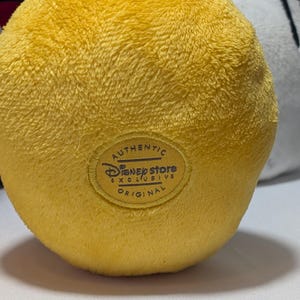 May include: Close-up of a plush yellow toy with a textured surface. The toy has a circular patch with the text "Authentic Disney Store Exclusive Original". Other plush toys are visible in the background.