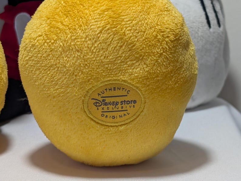 May include: Close-up of a plush yellow toy with a textured surface. The toy has a circular patch with the text "Authentic Disney Store Exclusive Original". Other plush toys are visible in the background.