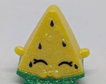 May include: A yellow, glittery, triangular toy resembling a watermelon slice. It has black seed details and a green, glittery rind. The toy has small, rounded yellow protrusions on either side.