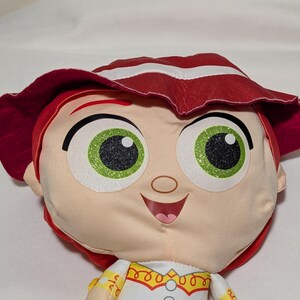 May include: A plush doll of a character with red hair and a matching hat with white stripes. The doll has large green eyes with black pupils and a smiling expression. The doll is wearing a white shirt with yellow and red accents.