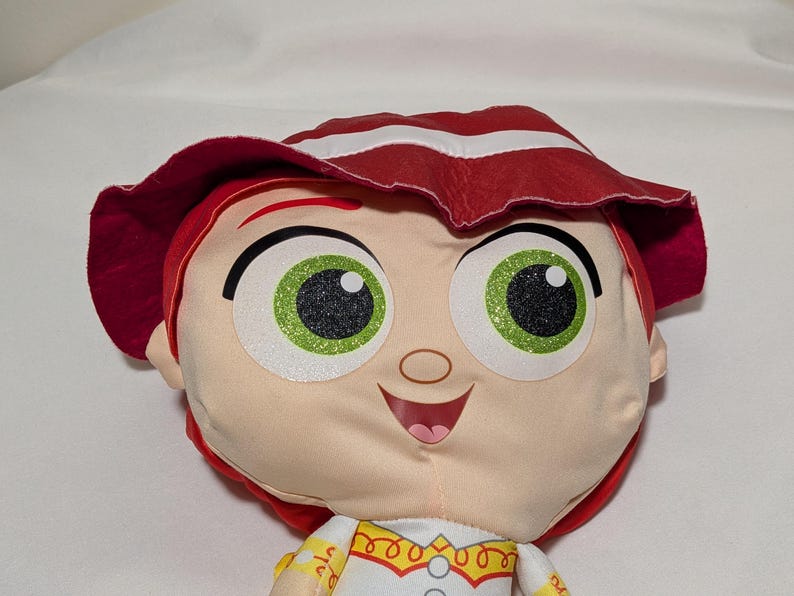 May include: A plush doll of a character with red hair and a matching hat with white stripes. The doll has large green eyes with black pupils and a smiling expression. The doll is wearing a white shirt with yellow and red accents.
