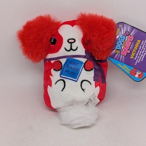 Könnte beinhalten: Ein Plüschtierhund mit roten Ohren und Akzenten, einem weißen Gesicht und einem kleinen weißen Schwanz. Das Spielzeug hat ein blaues Etikett mit der Aufschrift "EAT BARK PLAY". Ein lila Etikett mit den Worten "DOGS SQUIRIS" und "VICTORIA" ist angebracht.