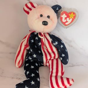 May include: A pink and white plush bear wearing a red, white and blue American flag pattern. The bear has black eyes and a black nose. The bear is holding a red heart-shaped tag with the word "ty" on it.
