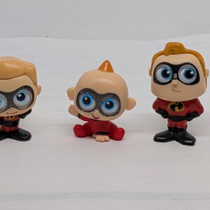 Può includere: Tre piccole statuette di personaggi de Gli Incredibili. Ogni statuetta ha un design a cartone animato con grandi occhi e un costume da supereroe. Le statuette sono di colore rosso, nero e marrone chiaro.