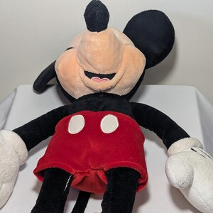 May include: A plush Mickey Mouse toy with a smiling expression. The toy features black ears, arms, and legs, a tan face, red shorts with white buttons, white gloves, and yellow shoes. The toy is designed to resemble the iconic Disney character.