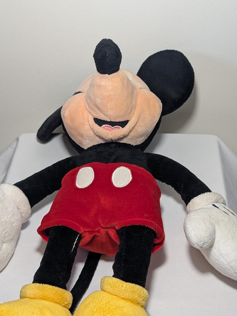 May include: A plush Mickey Mouse toy with a smiling expression. The toy features black ears, arms, and legs, a tan face, red shorts with white buttons, white gloves, and yellow shoes. The toy is designed to resemble the iconic Disney character.