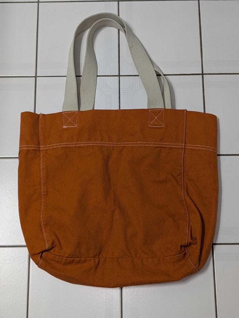 May include: A large, brown canvas tote bag with white straps. The bag has a simple design and is perfect for carrying everyday essentials.
