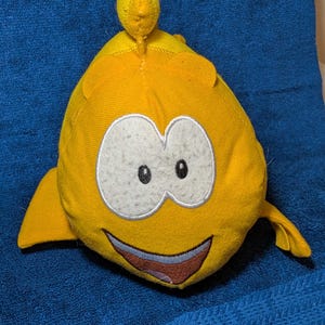 May include: A yellow plush toy shaped like a cartoon fish with large white eyes, a smiling mouth, and small fins. The toy has a small, round protrusion on top. The background is a blue fabric.
