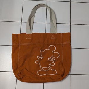 May include: A brown canvas tote bag with white straps and a white embroidered Mickey Mouse design.