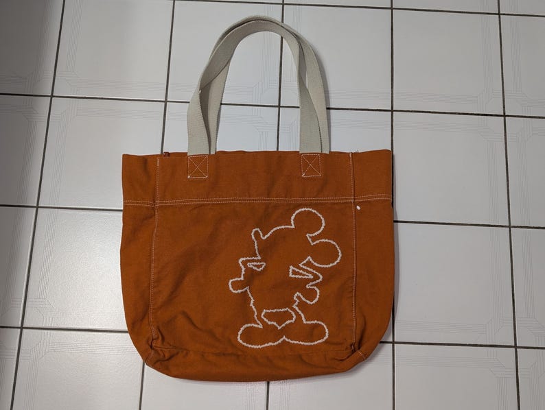 May include: A brown canvas tote bag with white straps and a white embroidered Mickey Mouse design.