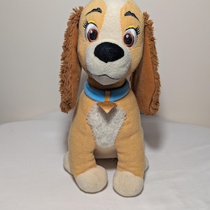 Può includere: Un peluche a forma di cane, con corpo beige, muso bianco e orecchie marroni. Il cane ha grandi occhi espressivi, un naso nero e un collare blu con un'etichetta dorata. Il giocattolo è seduto su una superficie bianca.