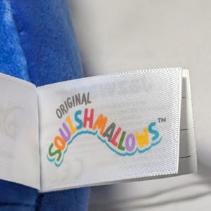 May include: Close-up of a white tag with the text "ORIGINAL SQUISHMALLOWS" in colorful, curved lettering. The tag is attached to a blue plush toy. The tag has a TM symbol.