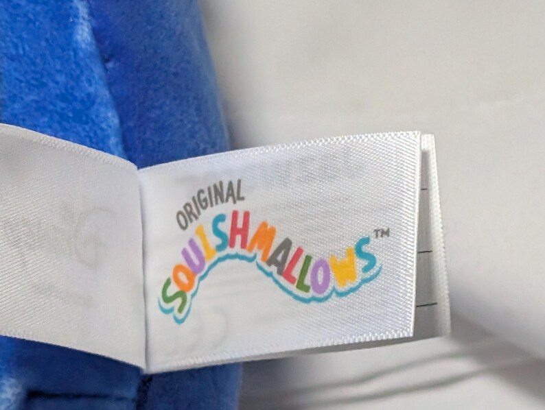 May include: Close-up of a white tag with the text "ORIGINAL SQUISHMALLOWS" in colorful, curved lettering. The tag is attached to a blue plush toy. The tag has a TM symbol.