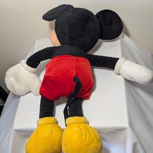 May include: A plush Mickey Mouse doll, viewed from the back. The doll features a black head and ears, a peach-colored face, black arms and legs, red shorts, white gloves, and yellow shoes. The doll's tail is visible.