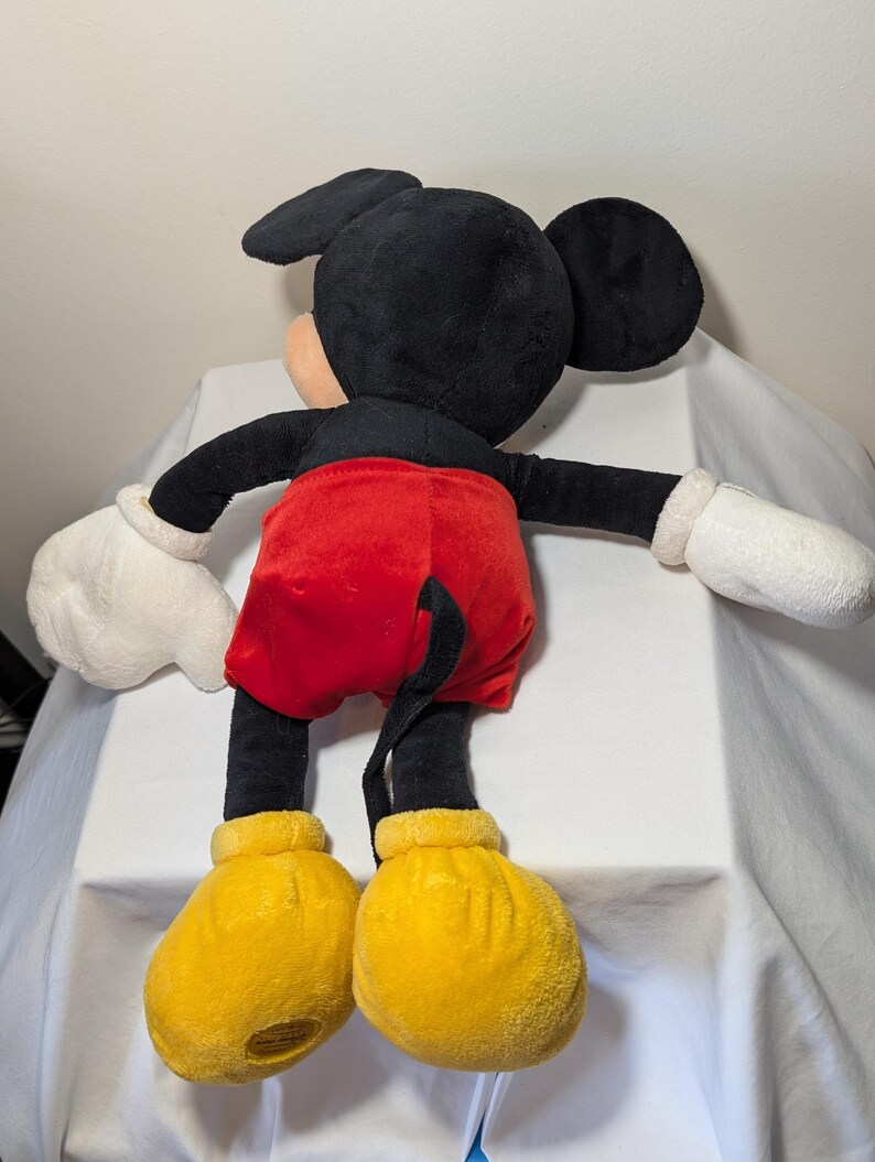 May include: A plush Mickey Mouse doll, viewed from the back. The doll features a black head and ears, a peach-colored face, black arms and legs, red shorts, white gloves, and yellow shoes. The doll's tail is visible.