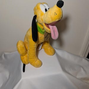 Puede incluir: Un peluche de Pluto, un perro amarillo con orejas y nariz negras. Tiene un collar verde y una lengua rosa. Una etiqueta con "Pluto" est&aacute; sujeta al collar. Personaje de Disney.