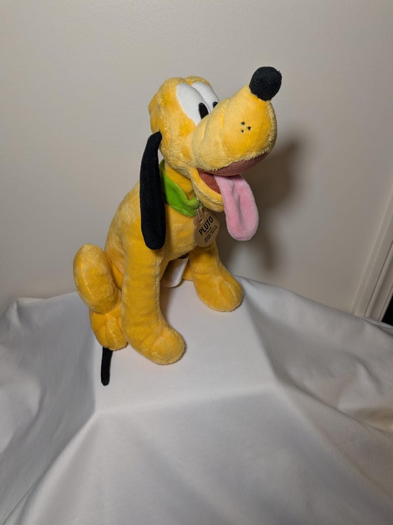Puede incluir: Un peluche de Pluto, un perro amarillo con orejas y nariz negras. Tiene un collar verde y una lengua rosa. Una etiqueta con "Pluto" est&aacute; sujeta al collar. Personaje de Disney.