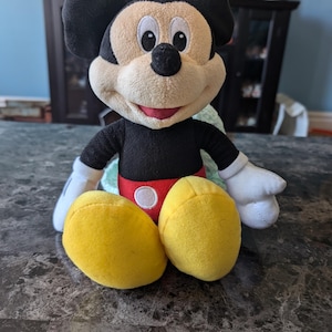 Puede incluir: Un juguete de peluche de Mickey Mouse con orejas negras, cuerpo negro, pantalones cortos rojos y zapatos amarillos. El juguete est&aacute; sentado en una mesa.