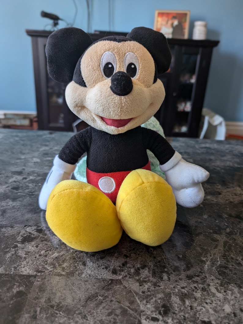 Puede incluir: Un juguete de peluche de Mickey Mouse con orejas negras, cuerpo negro, pantalones cortos rojos y zapatos amarillos. El juguete est&aacute; sentado en una mesa.