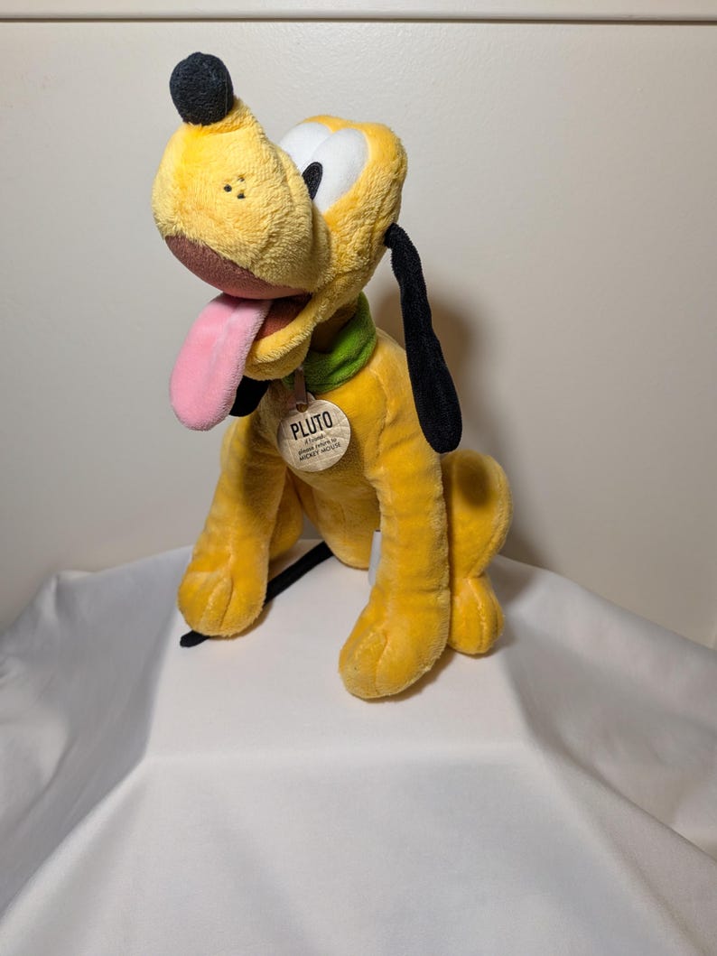 Puede incluir: Un peluche amarillo de Pluto con nariz, orejas y cola negras. El perro tiene una lengua rosa, un collar verde y una etiqueta que dice "Pluto". El juguete est&aacute; sobre una superficie blanca.