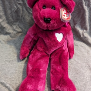 May include: A pink Beanie Baby plush bear with a white heart on its chest. The bear has a black nose and black eyes. The bear is wearing a red tag with the Beanie Baby logo.