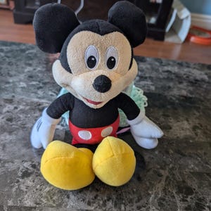 Puede incluir: Un juguete de peluche Mickey Mouse negro y amarillo sentado en un suelo de baldosas grises y blancas. El juguete lleva pantalones cortos rojos con lunares blancos y una gran sonrisa.