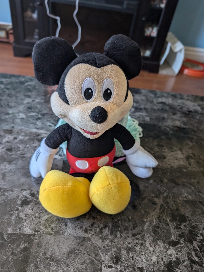 Puede incluir: Un juguete de peluche Mickey Mouse negro y amarillo sentado en un suelo de baldosas grises y blancas. El juguete lleva pantalones cortos rojos con lunares blancos y una gran sonrisa.