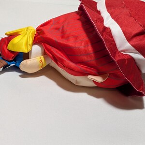 May include: A plush doll with a red dress and white trim, a yellow bow, and brown shoes. The doll is lying on its side. The design suggests a character from a children's story or cartoon. The doll's features are clearly visible.