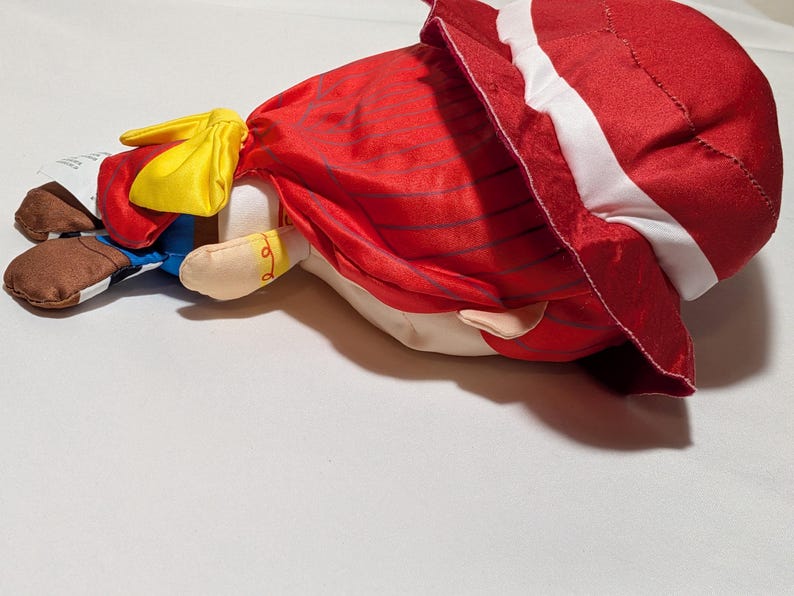 May include: A plush doll with a red dress and white trim, a yellow bow, and brown shoes. The doll is lying on its side. The design suggests a character from a children's story or cartoon. The doll's features are clearly visible.