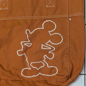 May include: A brown canvas tote bag with white stitching. The bag features a white embroidered outline of Mickey Mouse.