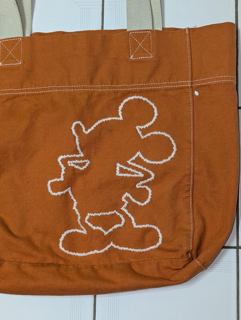 May include: A brown canvas tote bag with white stitching. The bag features a white embroidered outline of Mickey Mouse.