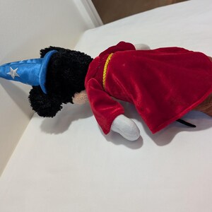 May include: A plush toy of a mouse dressed as a sorcerer. The mouse has black fur, white gloves, and brown shoes. It wears a red velvet robe with gold trim and a blue wizard hat with a silver star.