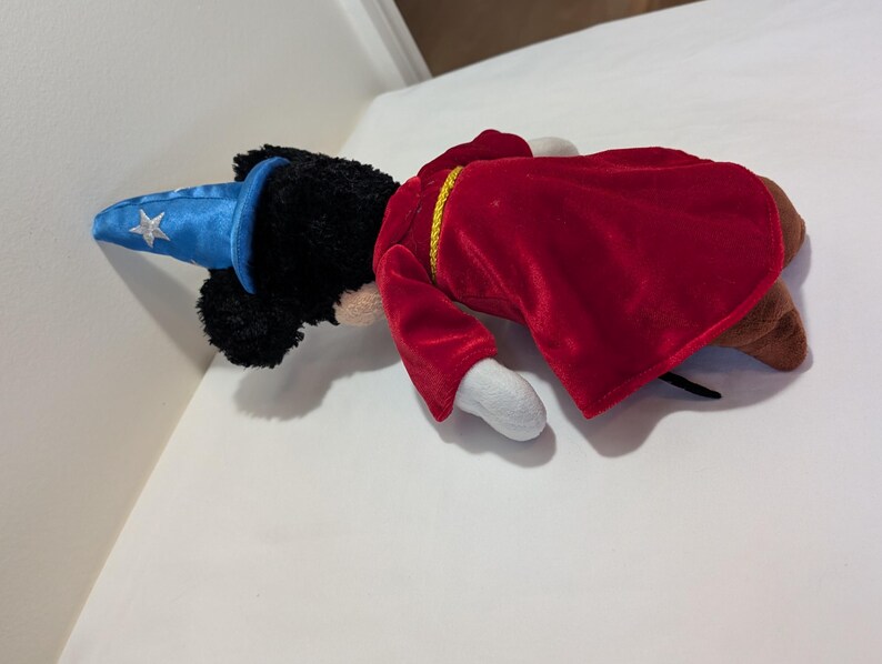 May include: A plush toy of a mouse dressed as a sorcerer. The mouse has black fur, white gloves, and brown shoes. It wears a red velvet robe with gold trim and a blue wizard hat with a silver star.