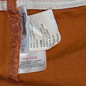 May include: A brown canvas bag with a white and orange label that reads "Disneyland Resort" and includes care instructions in English, French, Spanish, and Chinese. The label also includes a warning about a potential entanglement hazard.