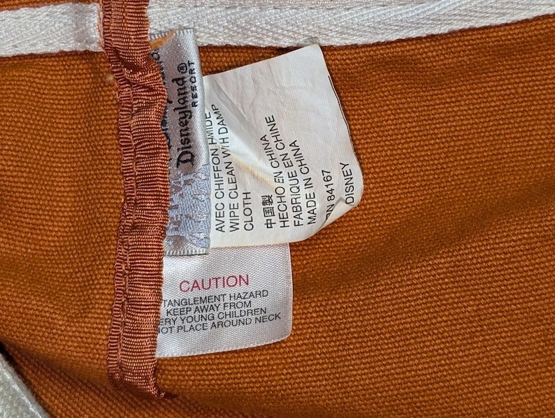 May include: A brown canvas bag with a white and orange label that reads "Disneyland Resort" and includes care instructions in English, French, Spanish, and Chinese. The label also includes a warning about a potential entanglement hazard.