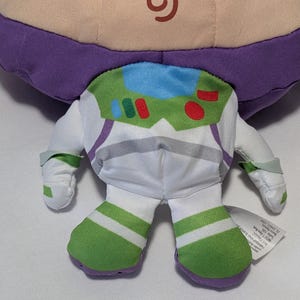 May include: A plush toy of Buzz Lightyear from Toy Story, featuring a purple head, white and green spacesuit, and green and white striped boots. The toy has a green and white chest plate with red and blue accents.