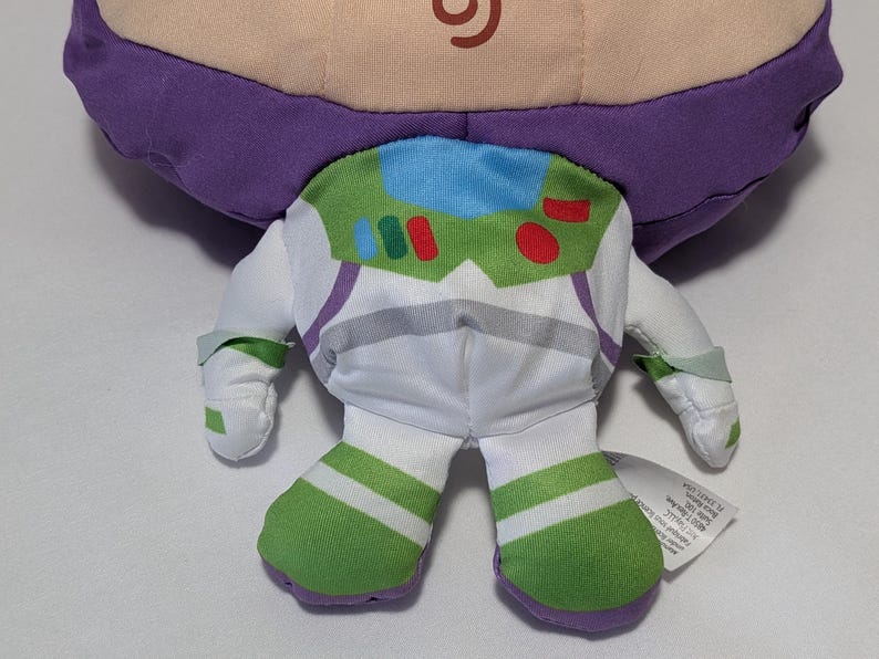 May include: A plush toy of Buzz Lightyear from Toy Story, featuring a purple head, white and green spacesuit, and green and white striped boots. The toy has a green and white chest plate with red and blue accents.