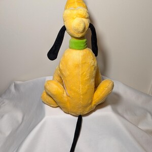 Puede incluir: Un peluche de Pluto, un personaje de perro amarillo con orejas y cola negras, un collar verde y un copete negro. El juguete est&aacute; sentado de espaldas al espectador, mostrando sus caracter&iacute;sticas.