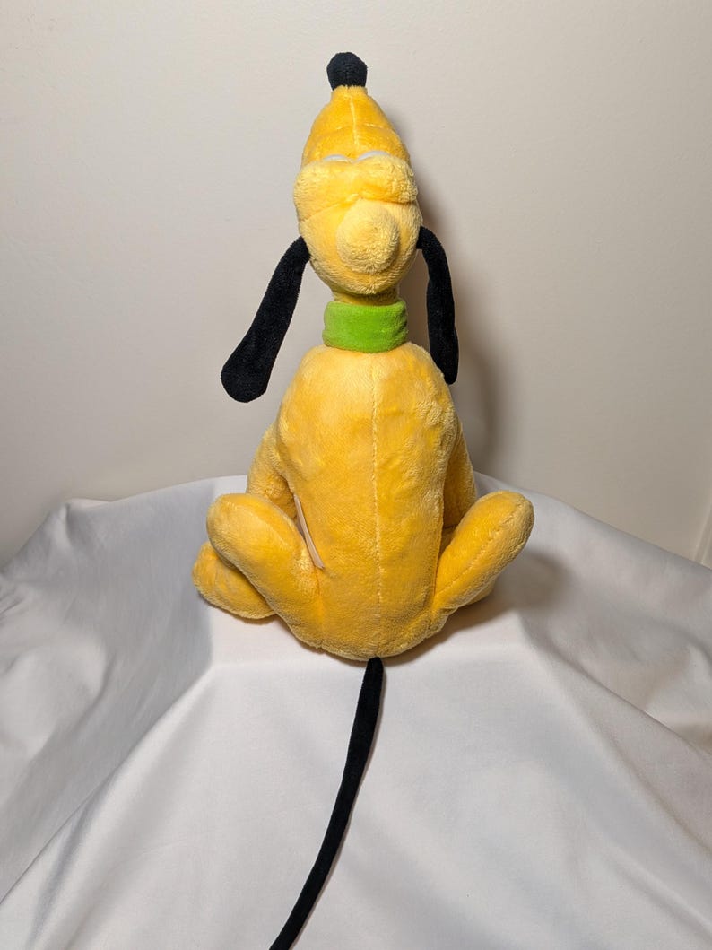 Puede incluir: Un peluche de Pluto, un personaje de perro amarillo con orejas y cola negras, un collar verde y un copete negro. El juguete est&aacute; sentado de espaldas al espectador, mostrando sus caracter&iacute;sticas.